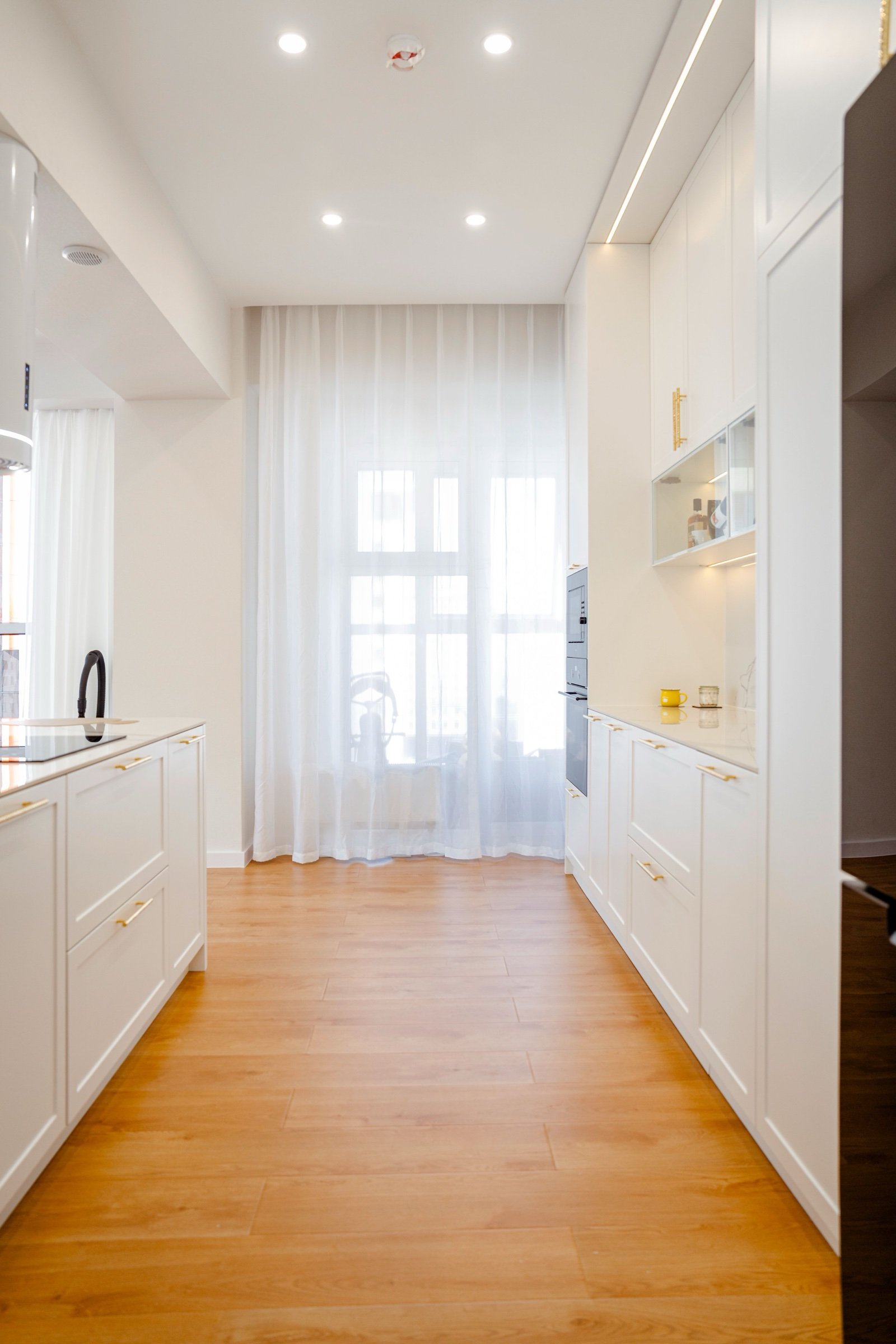 Renovated galley kitchen with white cabinets, oak-look flooring and daylight through sheer curtains